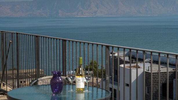 Ein Balkon mit einem Glastisch, einer Weinflasche und zwei Gläsern. Man sieht Wasser und sanfte Hügel in der Ferne.