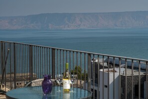 A balcony with a glass table, a bottle of wine, and two glasses. The view includes a body of water and distant hills. - Nino Lighthouse (Tiberias)