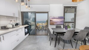 Eine moderne Küche mit weißen Schränken, ein Essbereich mit grauen Stühlen, ein großes Fenster mit Blick auf die Stadt und ein Balkon mit Tisch und Stühlen.