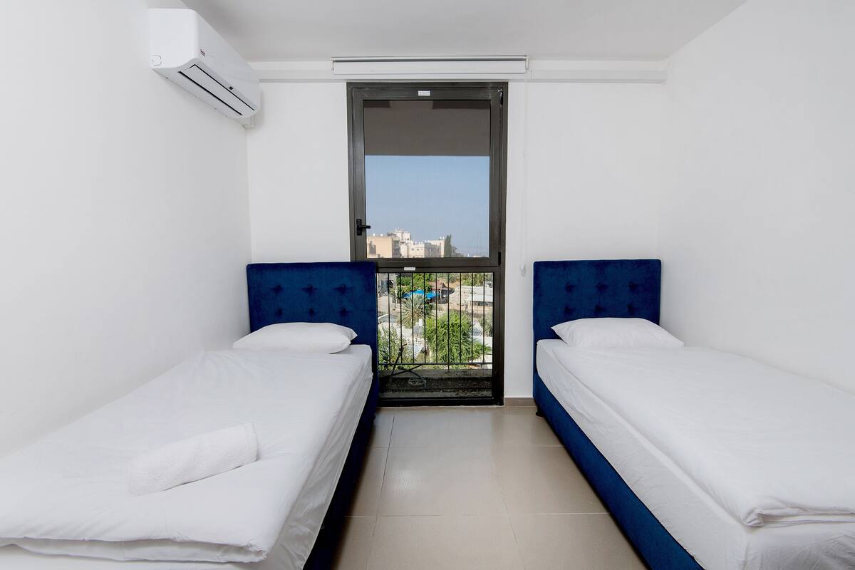 Una habitación pequeña y limpia con dos camas individuales, una ventana con vistas a edificios y un equipo de aire acondicionado en la pared.