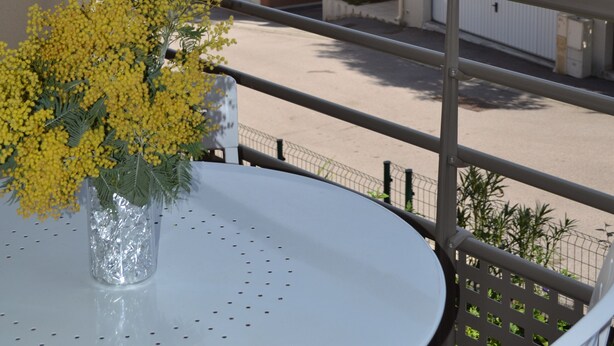 A balcony with a white table and a vase of yellow flowers, overlooking a residential area with red-tiled roofs and palm trees. - T3, 60 m2, small building with swimming pool, 1st floor, separate entrance (Collioure)