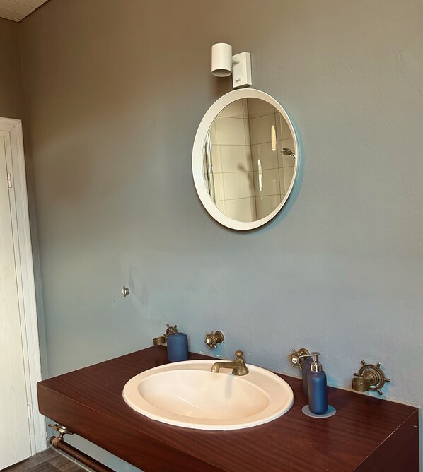 Casa de banho com um espelho redondo, uma pia com duas torneiras e um rolo de papel higiénico na parede.