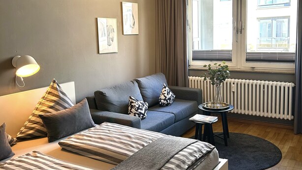 Un dormitorio moderno con una cama grande, un sofá y una pequeña mesita de noche. Hay una ventana con cortinas, un radiador y una planta sobre la mesita.