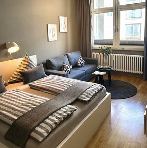 Quarto moderno com uma cama grande, um sofá e uma pequena mesa lateral. Há uma janela com cortinas, um radiador e uma planta na mesa lateral.