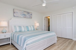 Une chambre à coucher comprenant un lit, deux tables de chevet avec des lampes, un ventilateur de plafond, une porte donnant sur une autre pièce et un placard avec des portes à persiennes.