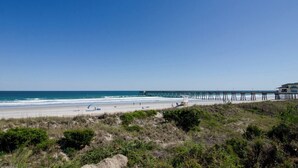 Una playa con un muelle que se adentra en el océano, arena y algo de vegetación al frente.