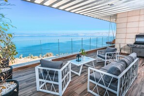 Patio esterno moderno con mobili bianchi, una griglia e vista sul mare.