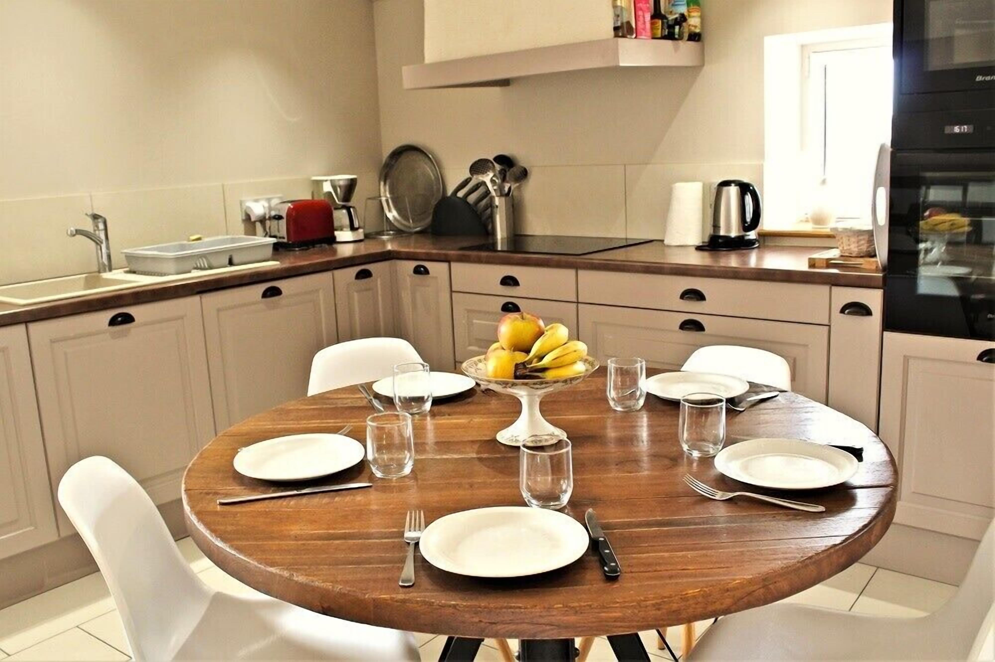 Una cocina con una mesa redonda de madera preparada para cuatro personas, sillas blancas, un frutero con plátanos y una cafetera.