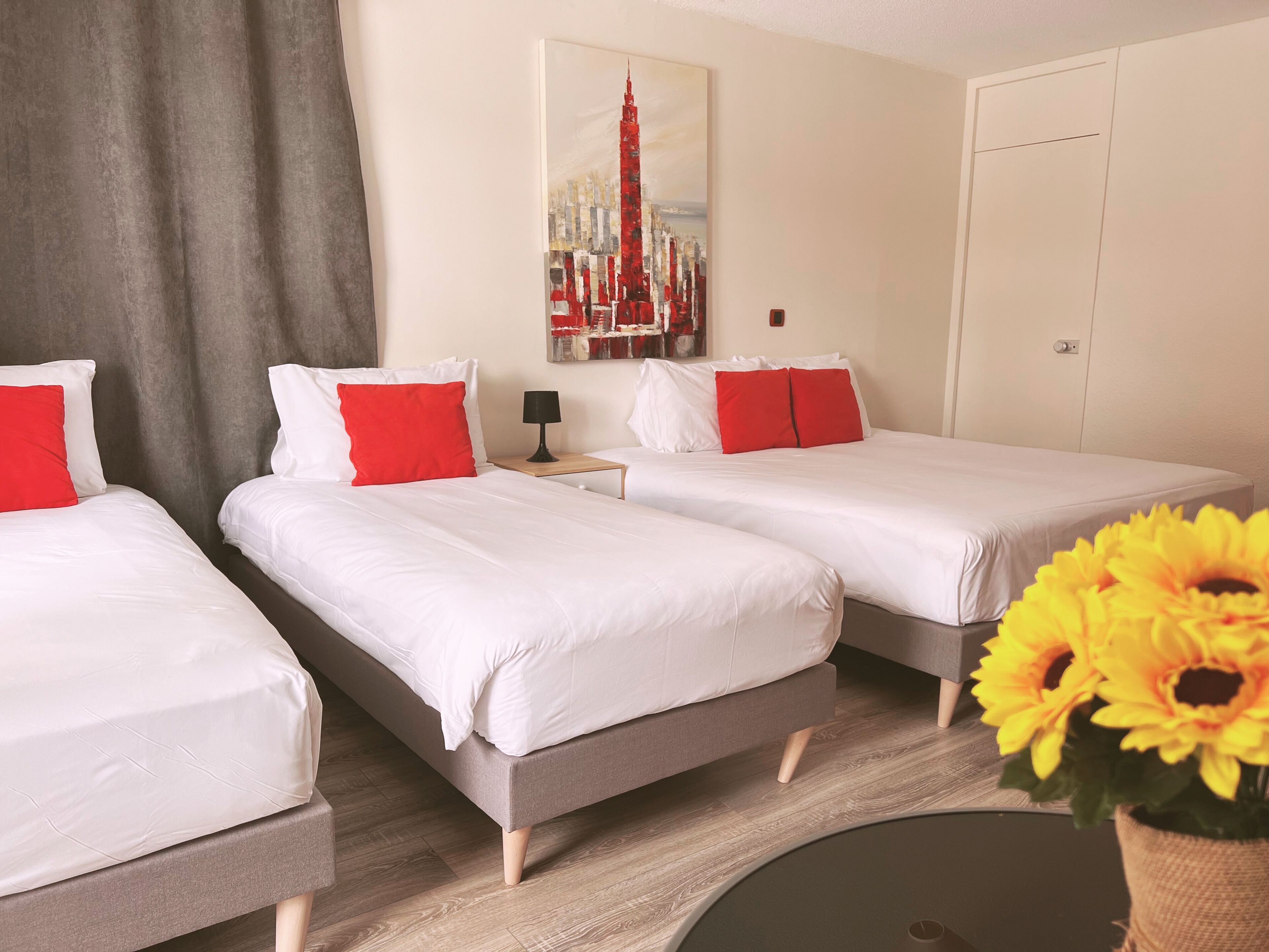 A hotel room with two beds, each with white bedding and red pillows, a nightstand with a lamp, a painting of a cityscape, and a vase with yellow flowers.
