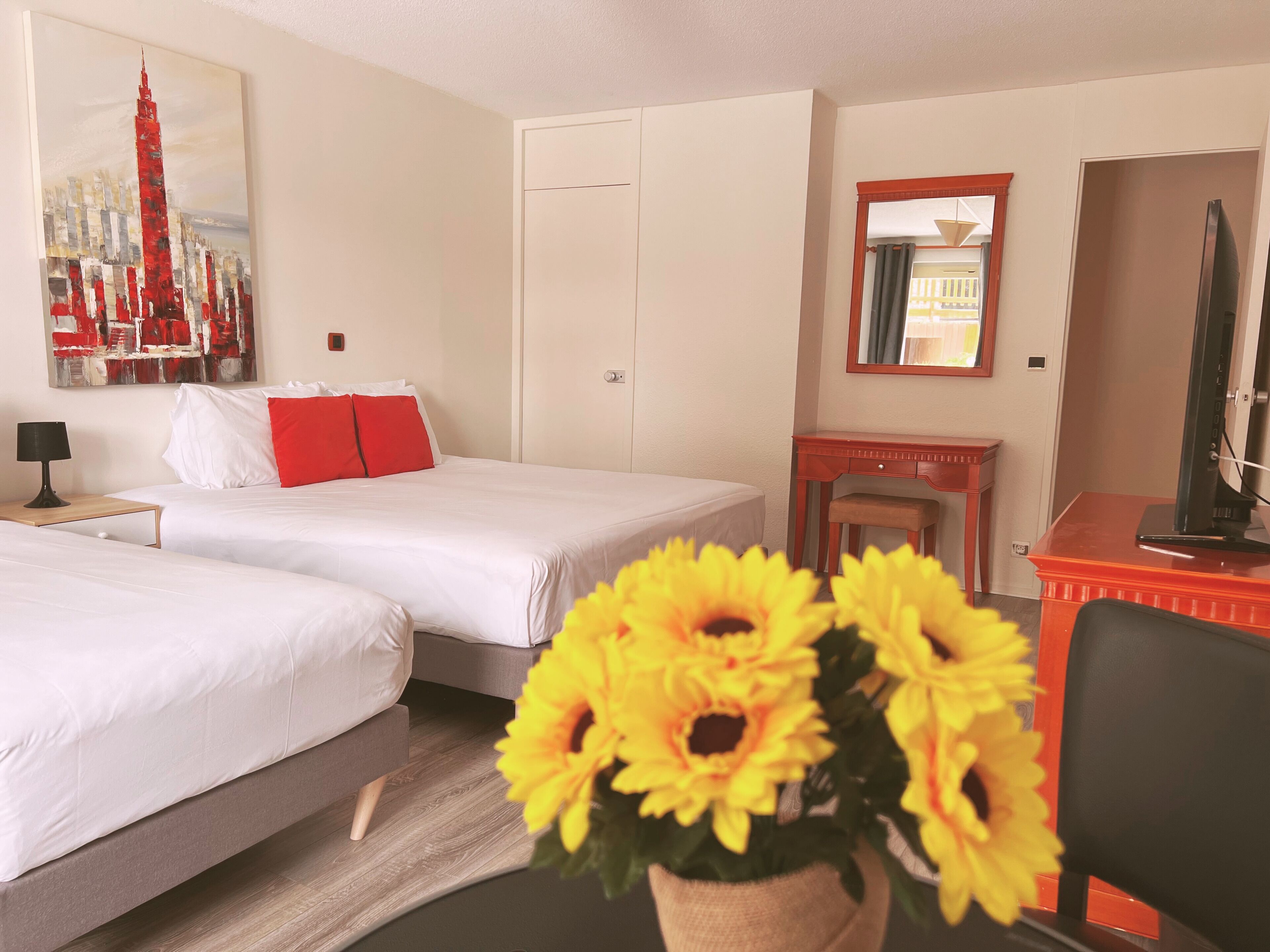 A hotel room with two beds, a desk, a chair, a TV, a mirror, and a painting of a cityscape.