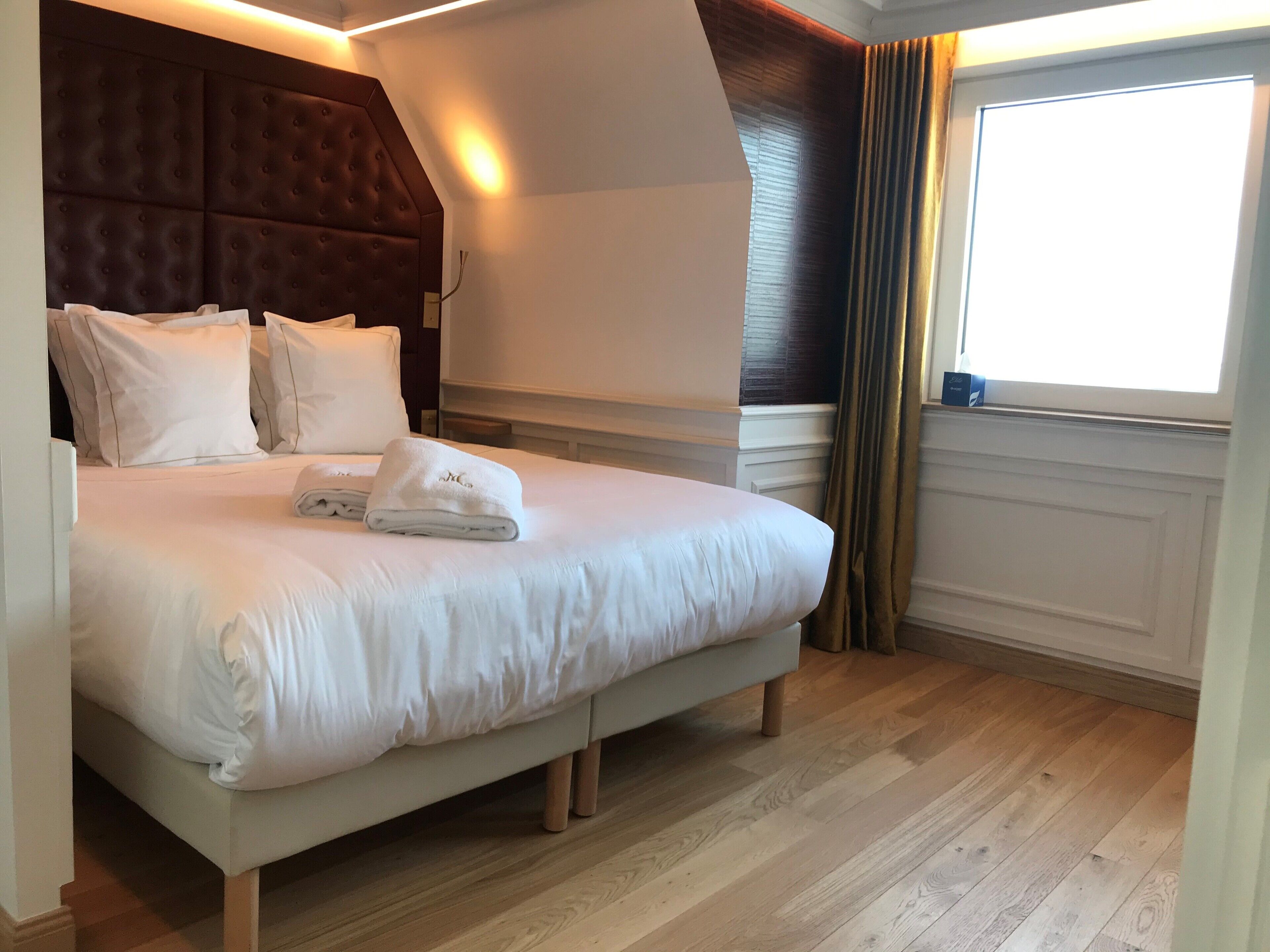 Ein ordentlich bezogenes Bett mit weißen Leinen und Kissen, ein gepolsterter Kopfteil und ein großes Fenster mit Vorhängen.