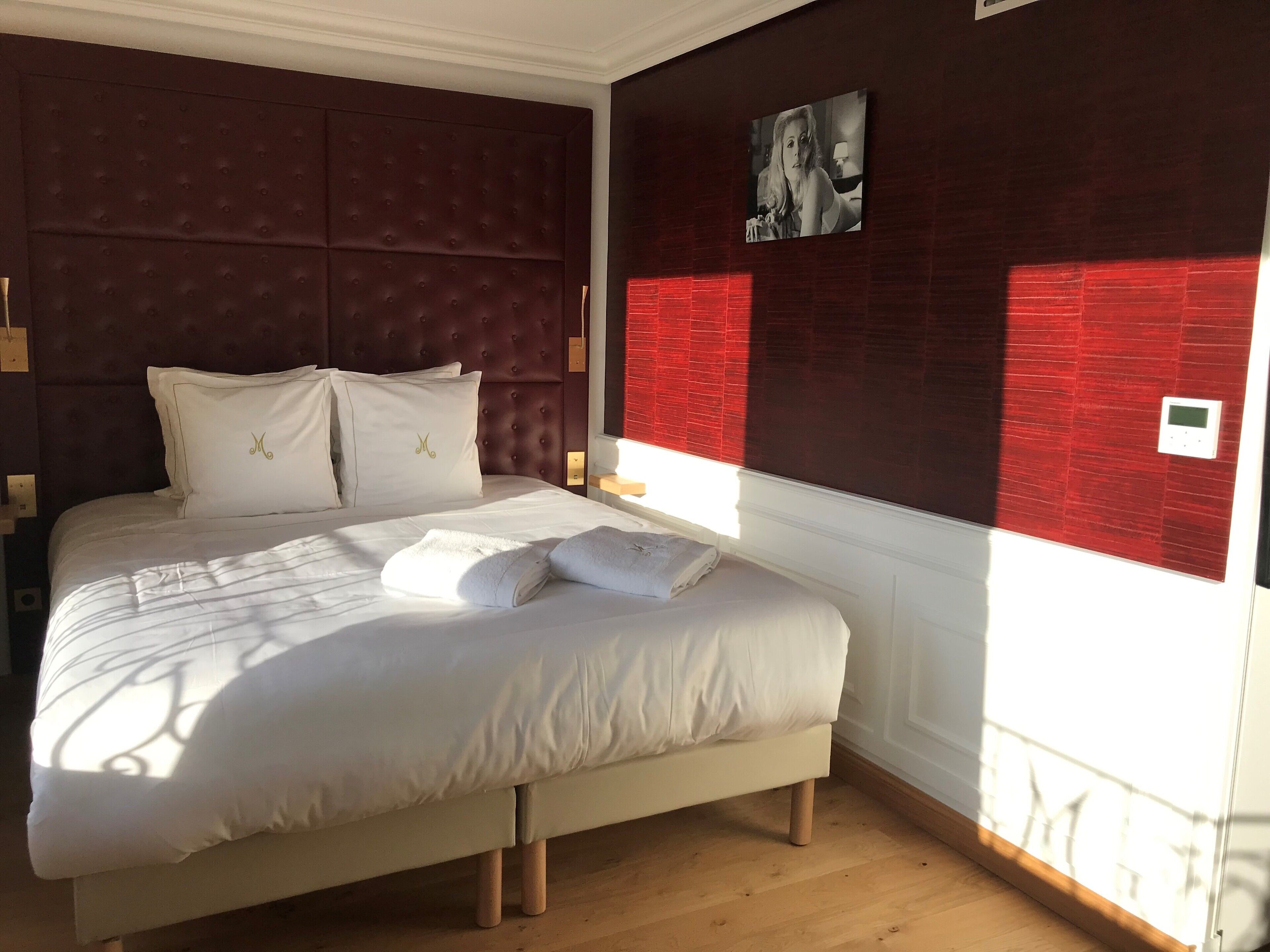 Cama bem arrumada com lençóis e almofadas brancos, cabeceira escura e parede de destaque vermelha com um quadro.