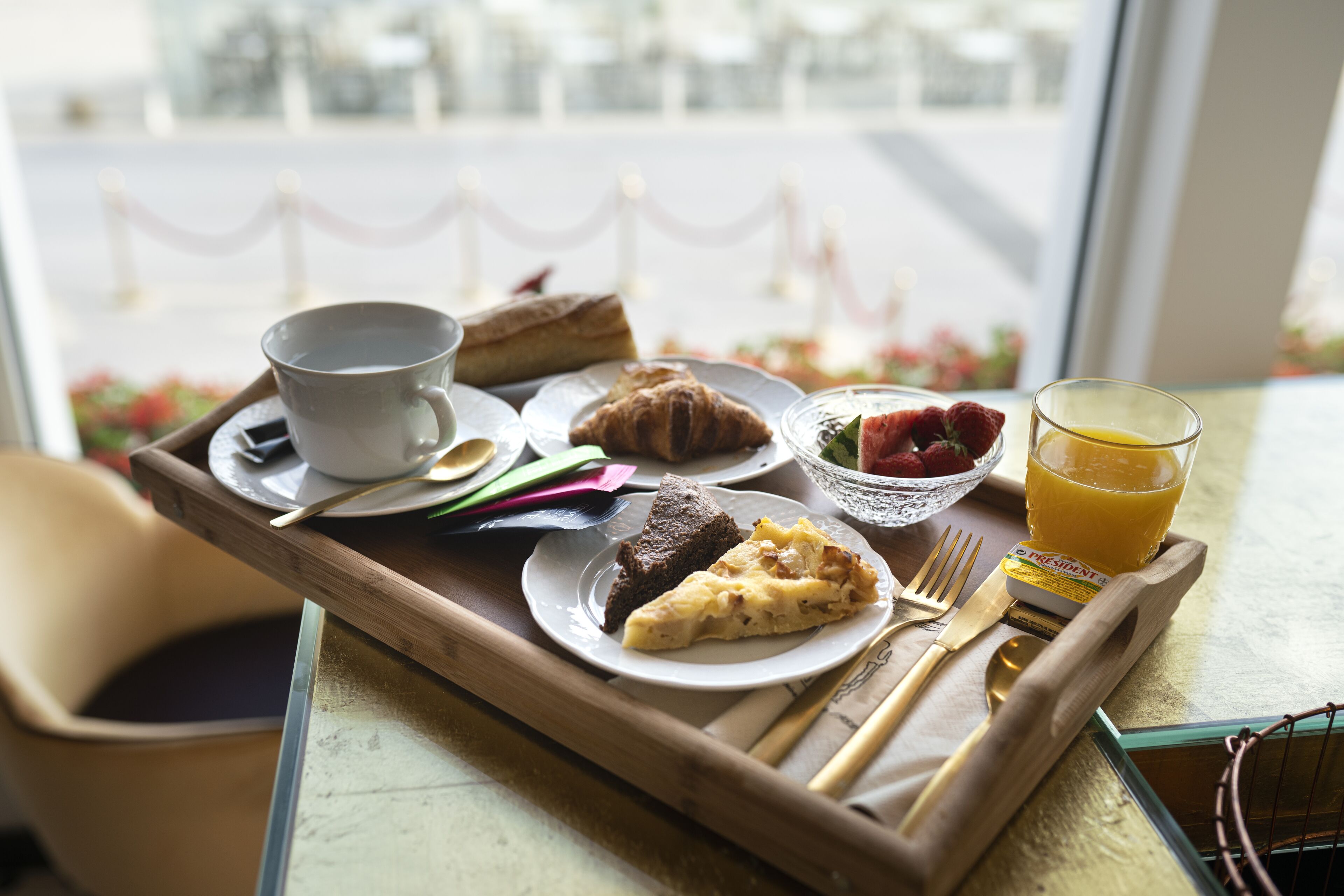 Un plateau de petit-déjeuner avec une tasse, un croissant, une part de gâteau, des fraises et un verre de jus d’orange, posé sur une table près d’une fenêtre.