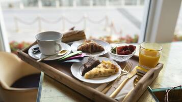 Un plateau de petit-déjeuner avec une tasse, un croissant, une part de gâteau, des fraises et un verre de jus d’orange, posé sur une table près d’une fenêtre.