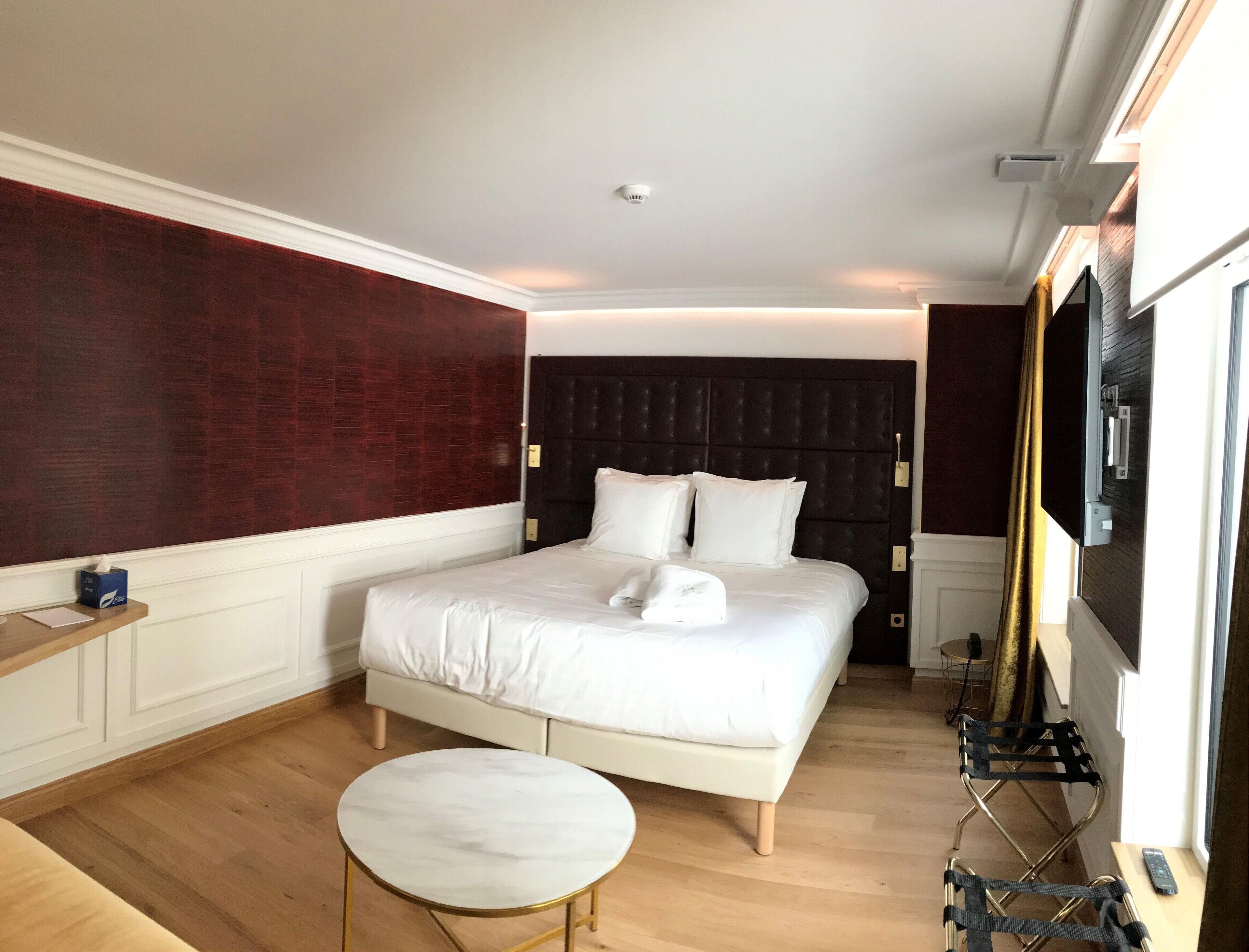 A hotel room with a large bed, a small round table, a TV, and a window with curtains.