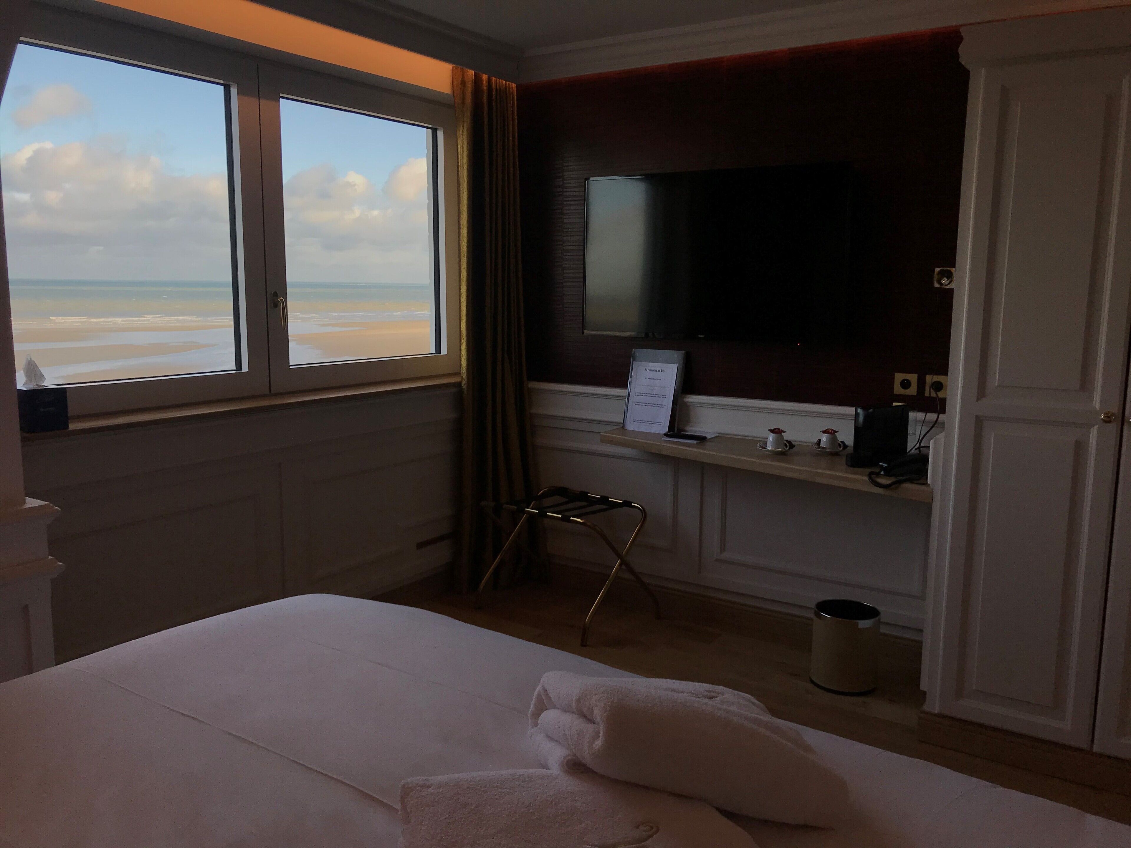 A hotel room with a bed, a television, a desk with a computer, a trash can, and a view of the beach.