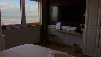 A hotel room with a bed, a television, a desk with a computer, a trash can, and a view of the beach.