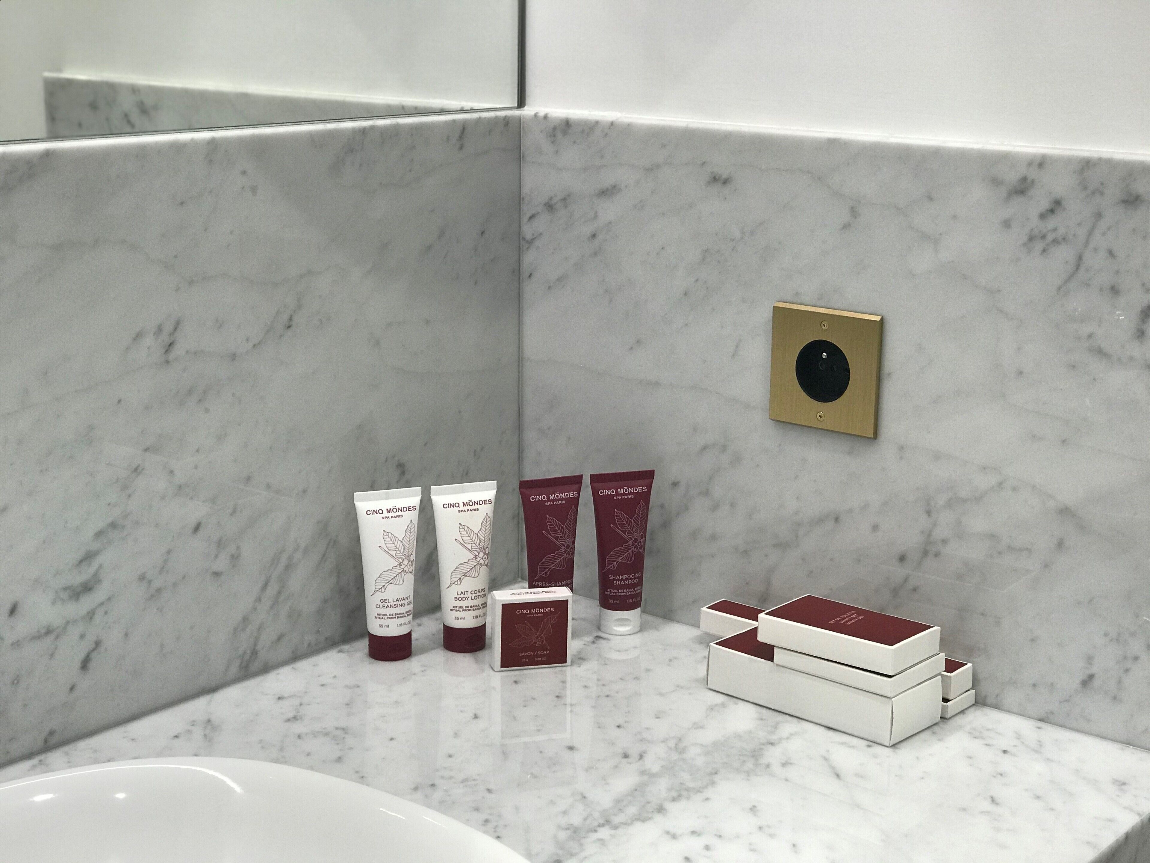 Une salle de bain avec des murs en marbre et un lavabo blanc. On y trouve trois tubes de lotion pour le corps de la marque Cinq Mondes et une boîte de shampoing de la même marque.