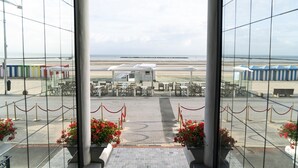 A view through glass doors to a beachside dining area with outdoor seating, a clear sky, and a sandy beach in the background. - Hotel Merveilleux Côté Mer (Dunkirk)