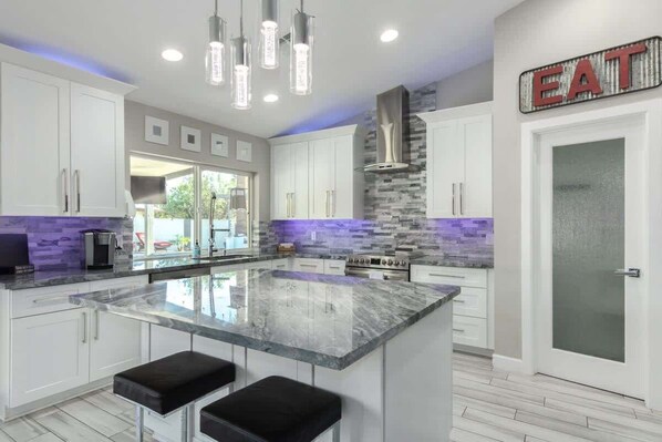 A modern kitchen with white cabinets, a large island with bar stools, a stone backsplash, and pendant lighting.