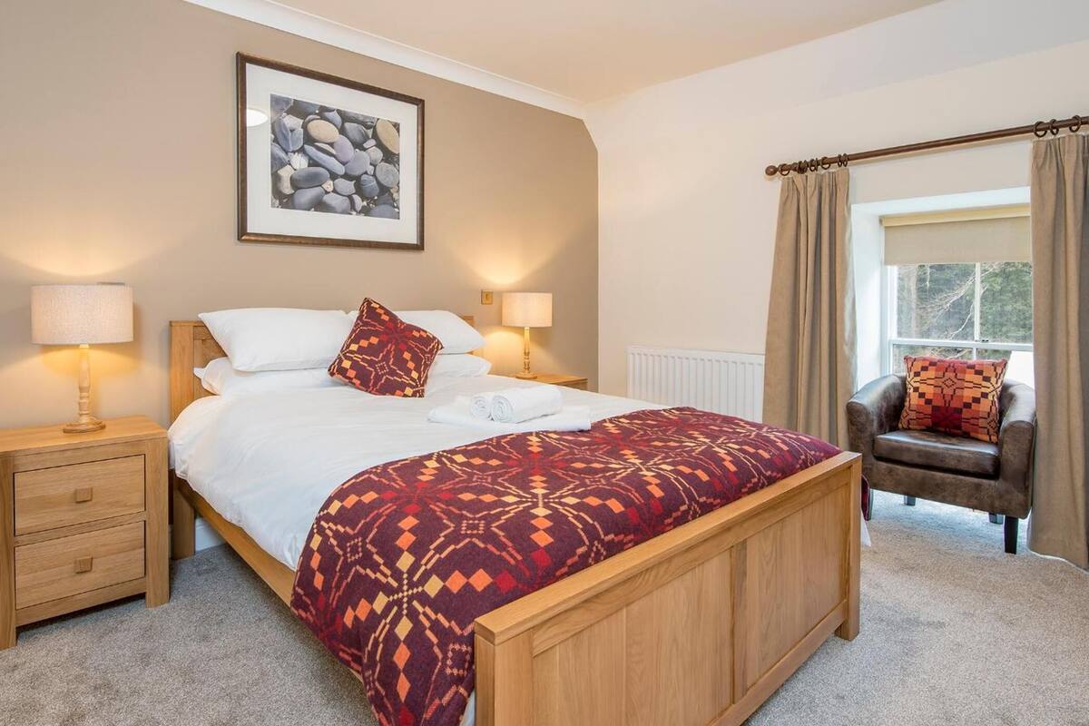 A bedroom with a bed, two bedside tables, a chair, a window with curtains, and a framed picture on the wall.