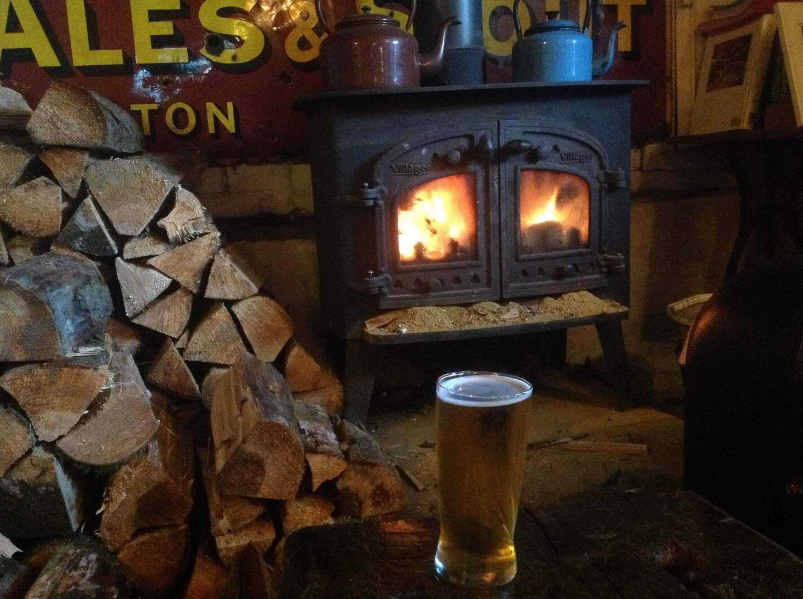 A wooden log stack, a wood-burning stove, and a glass of beer on a table.