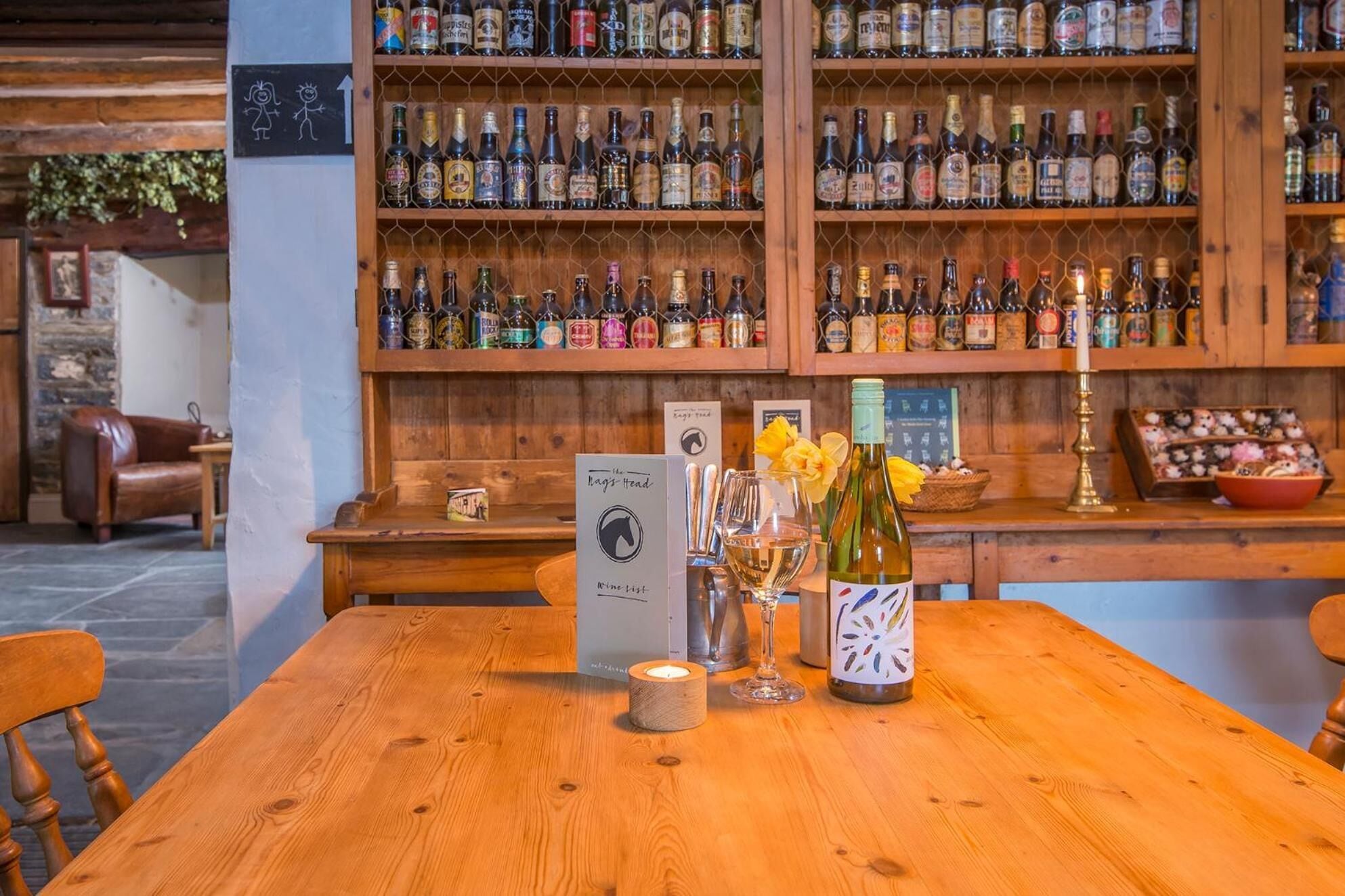 Un coin bar avec des étagères en bois remplies de différentes bouteilles d’alcool, une table en bois sur laquelle se trouvent une bougie, une bouteille de vin et un panneau indiquant le menu.