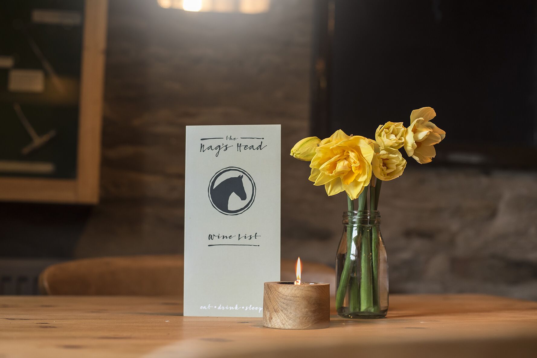 A wooden table with a candle, a sign reading "the Nag's Head Wine List," and a vase with yellow flowers.