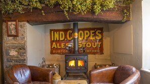 A cozy pub interior with leather chairs, a wooden table, and a sign for 'IND.COOPES ALES & STOUT' above a wood-burning stove. - The Nag's Head Inn (Boncath)