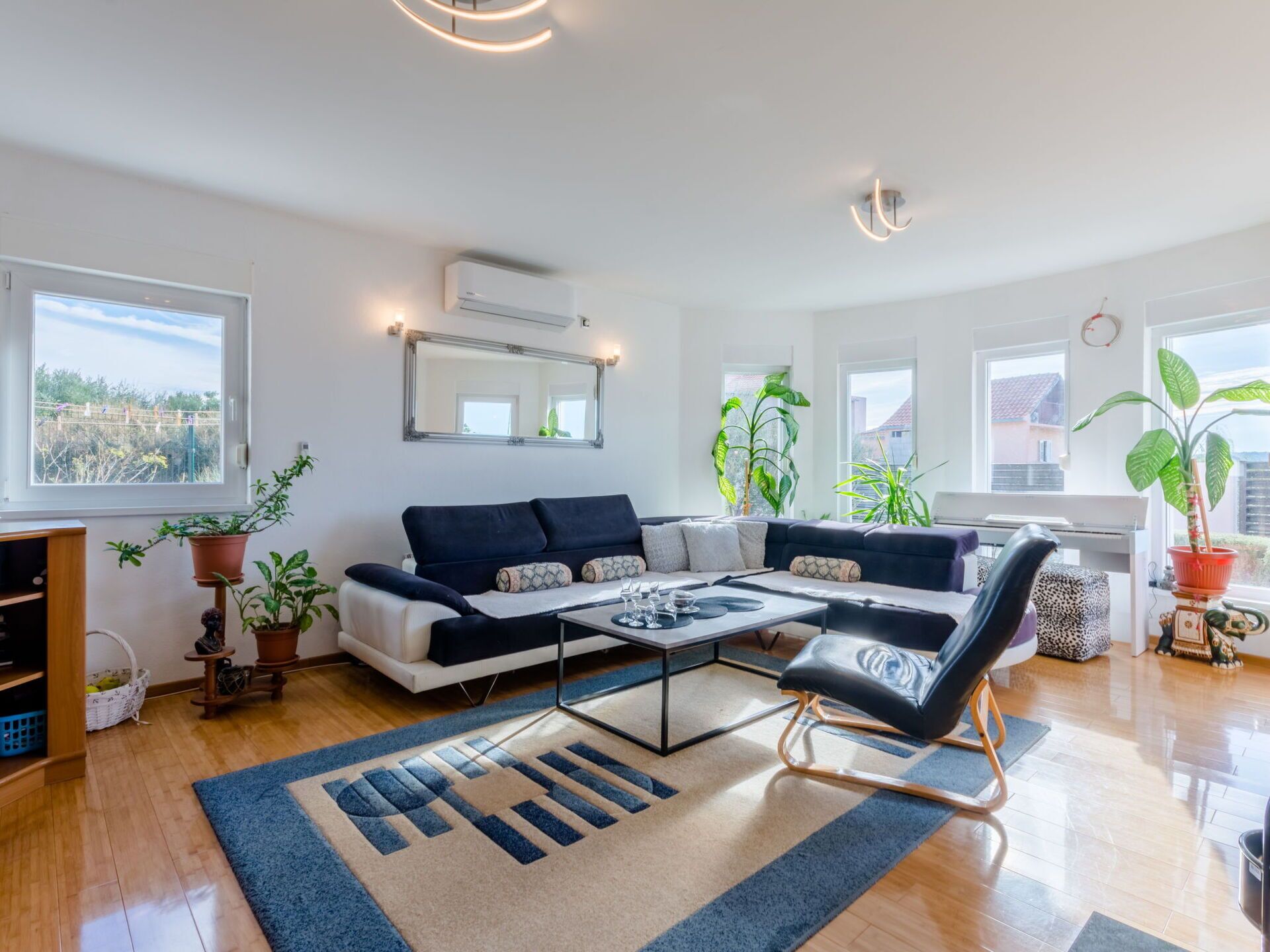 A modern living room with a sofa, coffee table, and a chair. There are potted plants, a mirror, and a window with a view.
