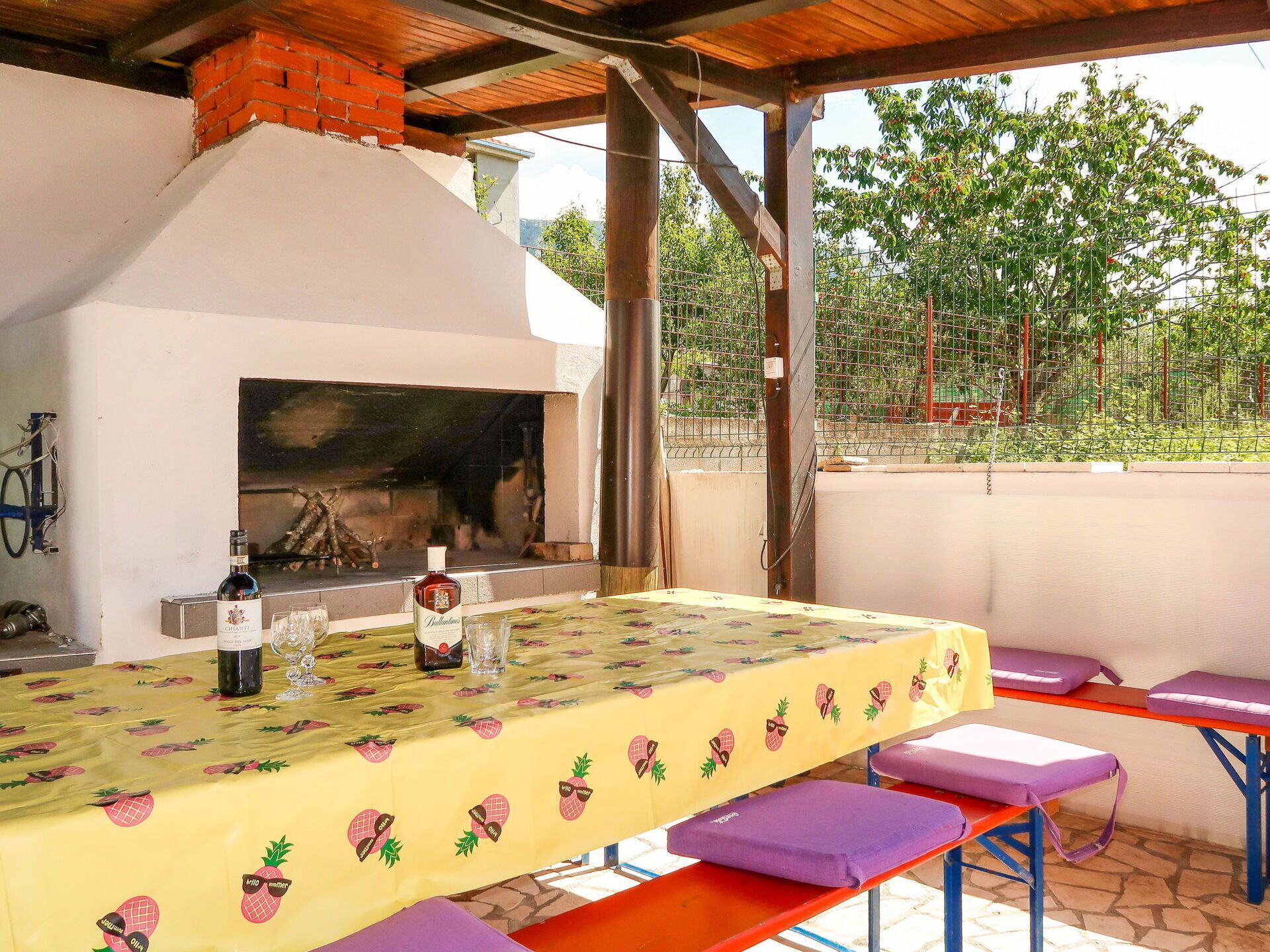 An outdoor patio with a barbecue grill, a table with a fruit-patterned tablecloth, and purple cushioned benches.