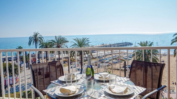 Een tafel voor twee gedekt met champagne, glazen en borden op een balkon met uitzicht op een strand met palmbomen en boten.
