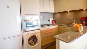 Une petite cuisine équipée d’un lave-linge, d’une machine à café et d’un panier rempli d’oranges posé sur le plan de travail.