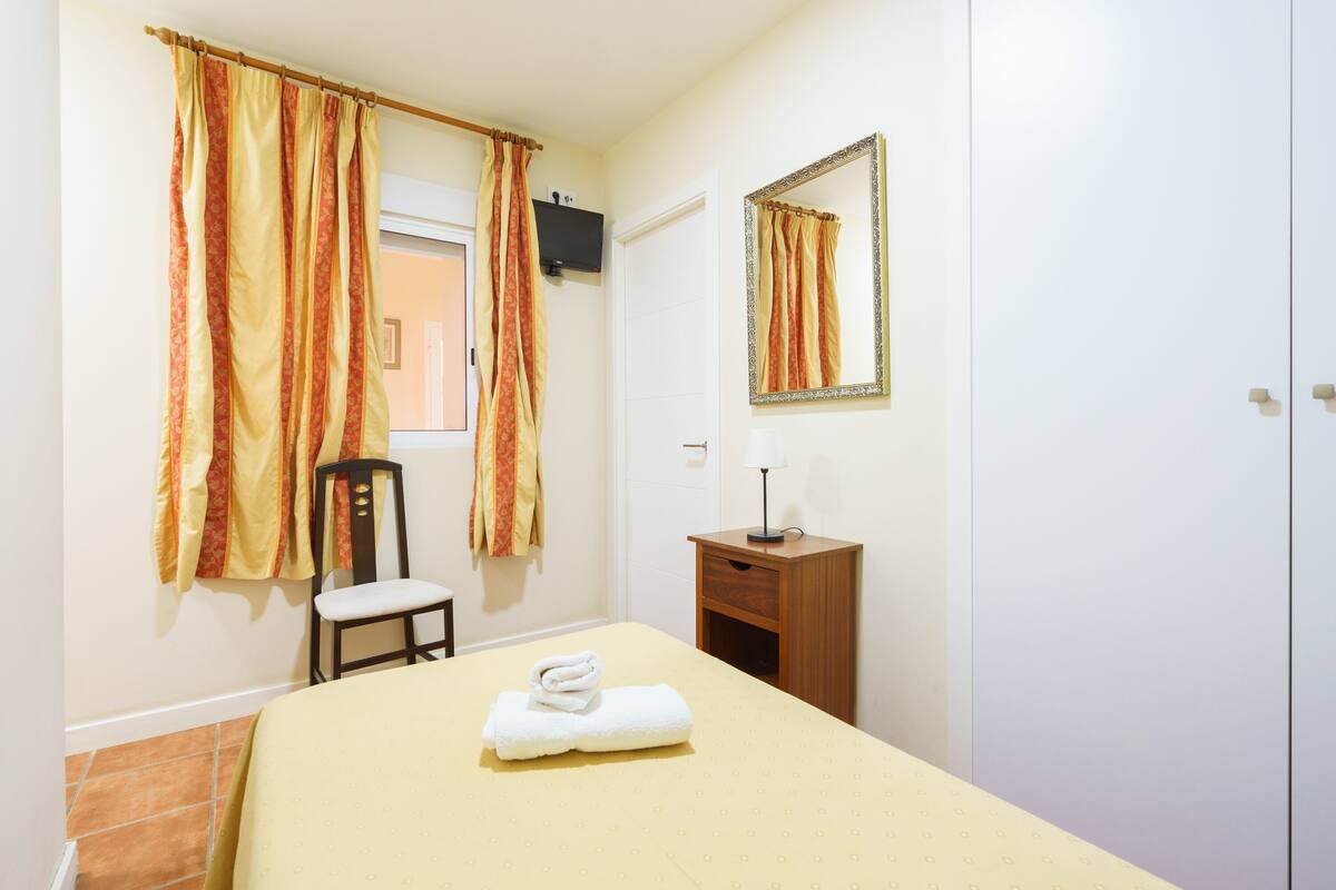 Una habitación de hotel con una cama, una silla, una mesita de noche de madera, un espejo y una ventana con cortinas.