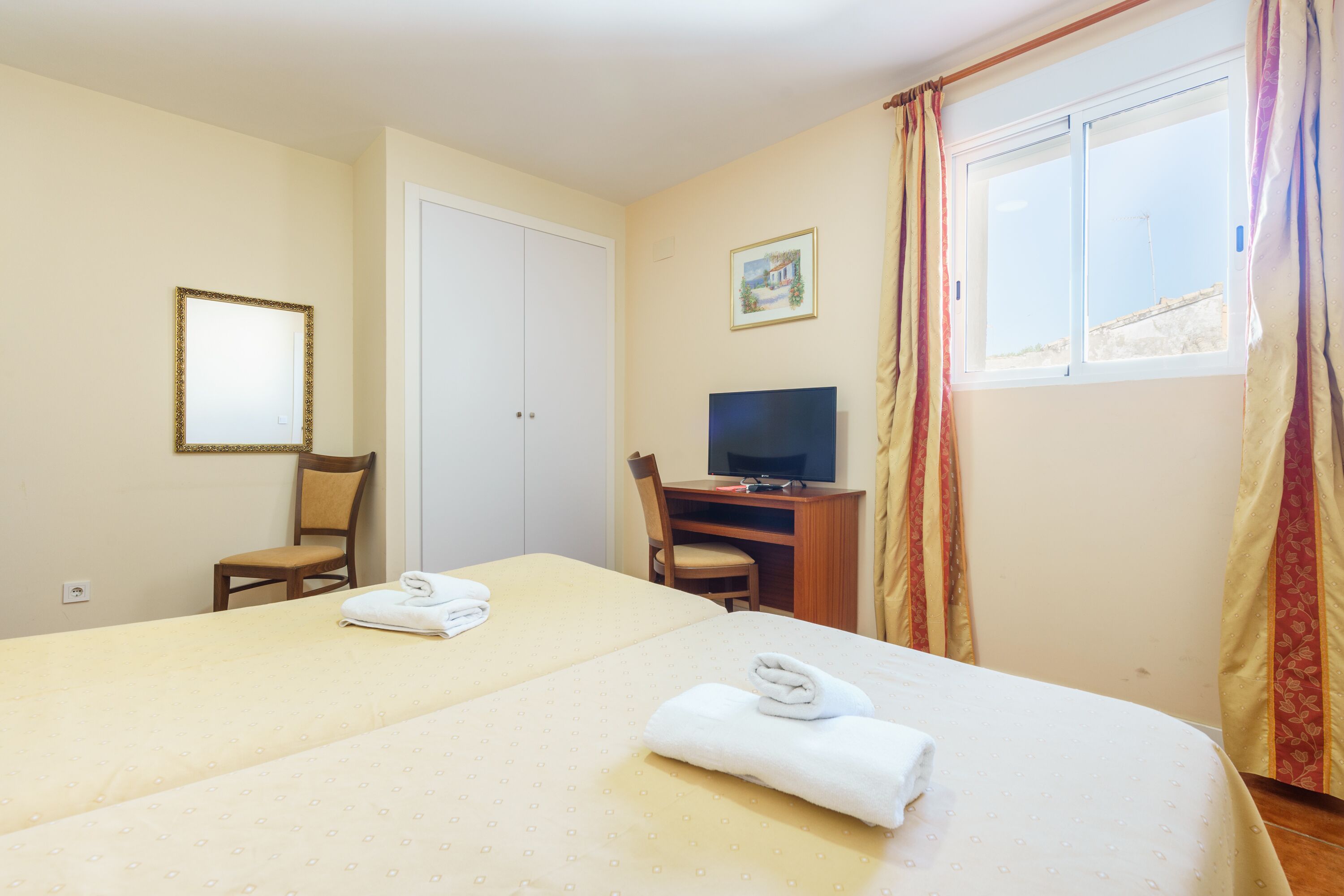 A hotel room with two beds, a TV, a desk, and a window with curtains.