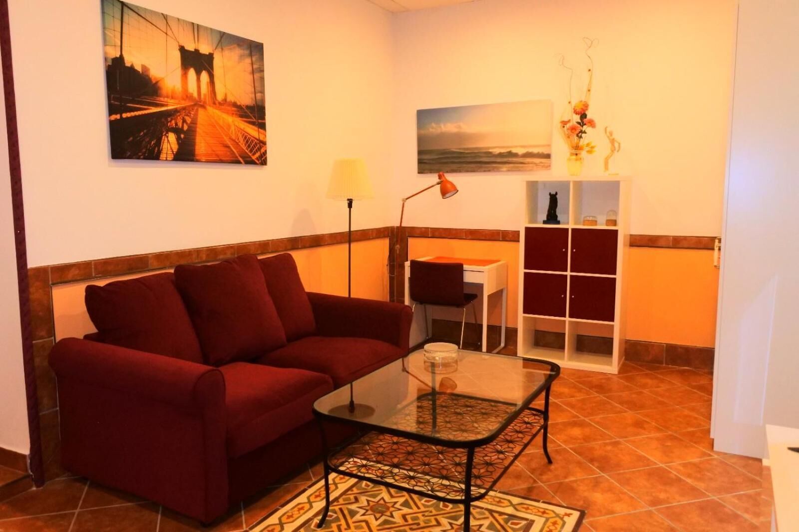 A living room with a red sofa, a glass coffee table, a lamp, and a painting of a bridge.