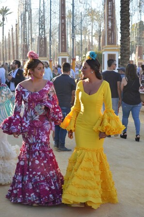 Dos mujeres con vestidos tradicionales caminando por un sendero arenoso, con palmeras y edificios al fondo.