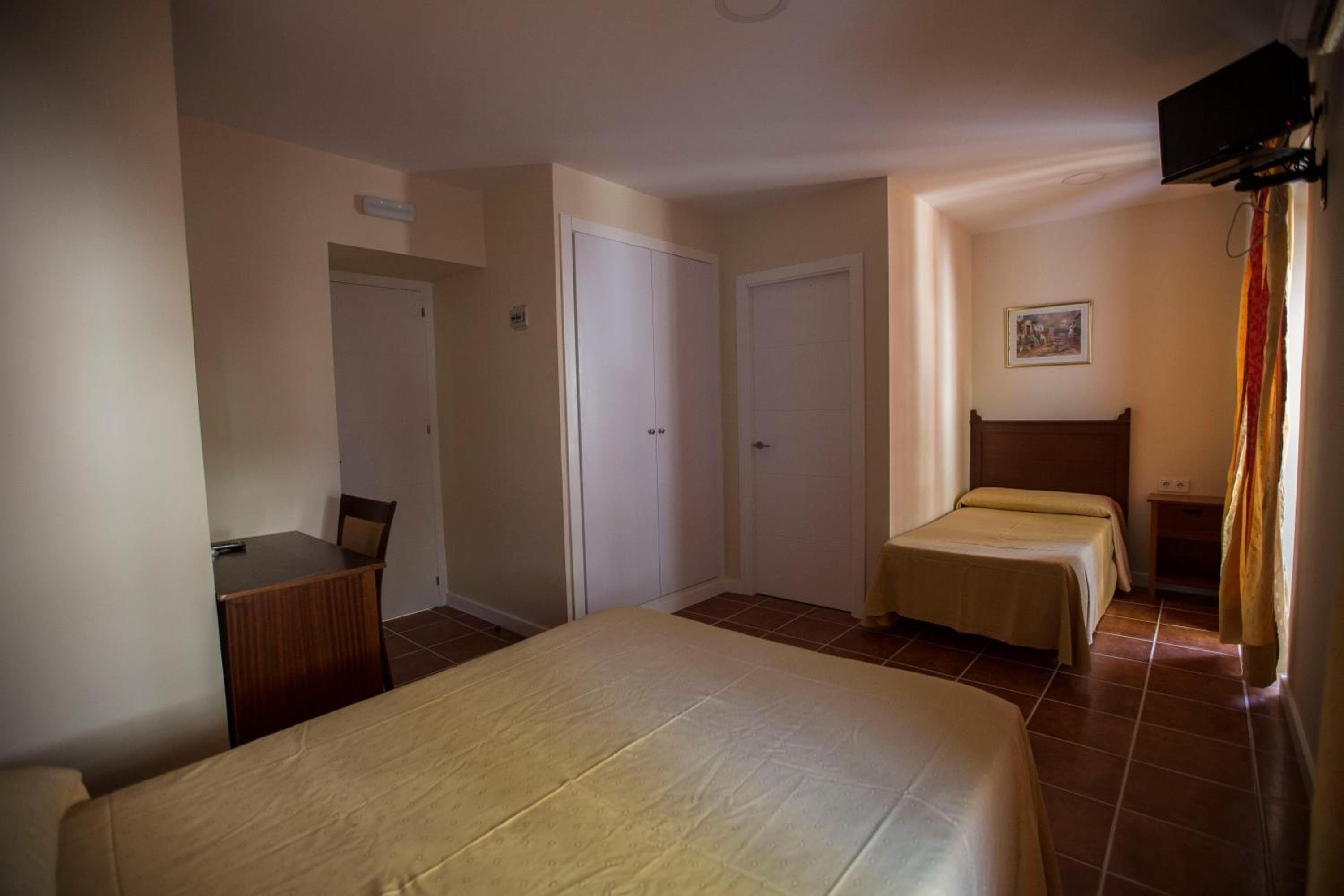 A hotel room with a large bed, two smaller beds, a desk with a chair, and a television mounted on the wall.