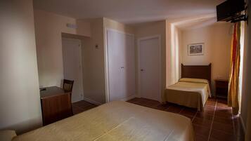 Habitación de hotel con una cama grande, dos camas individuales, un escritorio con silla y un televisor montado en la pared.