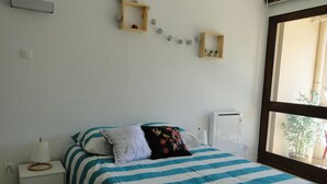 A bedroom with a bed, a nightstand, a wall-mounted shelf with decorative items, and a window with a view of greenery. - Comfortable studio with view of pond and Chateau (Chauffailles)