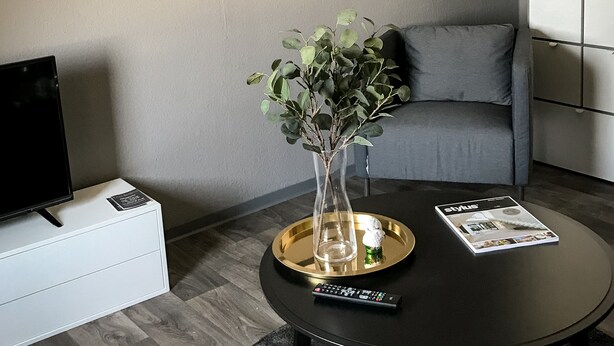 Un soggiorno moderno con un tavolino rotondo, una televisione su un mobile bianco, un quadro appeso al muro e un divano grigio.