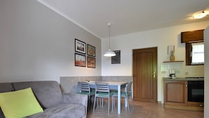 Un salon avec un canapé gris, une petite table en bois et un coin repas avec des chaises. On peut voir une cuisine avec un four et des placards.