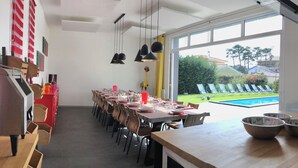 Un'area pranzo con un lungo tavolo apparecchiato per un pasto, una piscina visibile dalla finestra e un mobiletto rosso con contenitori portaoggetti.