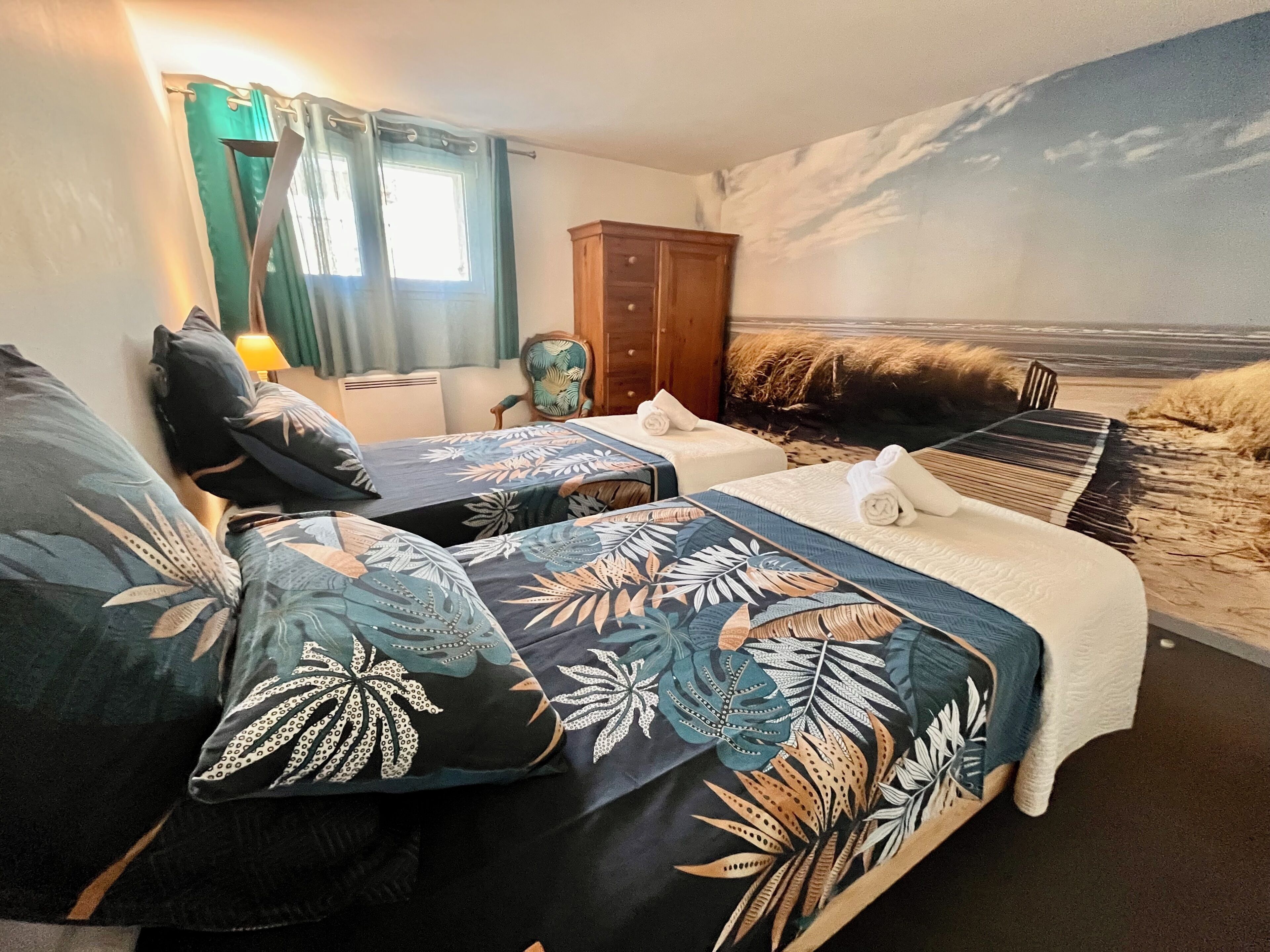 A room with two beds, a wooden wardrobe, a chair, and a beach mural on the wall.