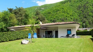 A small, single-story building with a tiled roof, a covered patio area with outdoor seating, and a view of a lush green mountain. - Lovely apartment in villa for 5 guests with WIFI, TV, terrace and pets allowed (Crone)