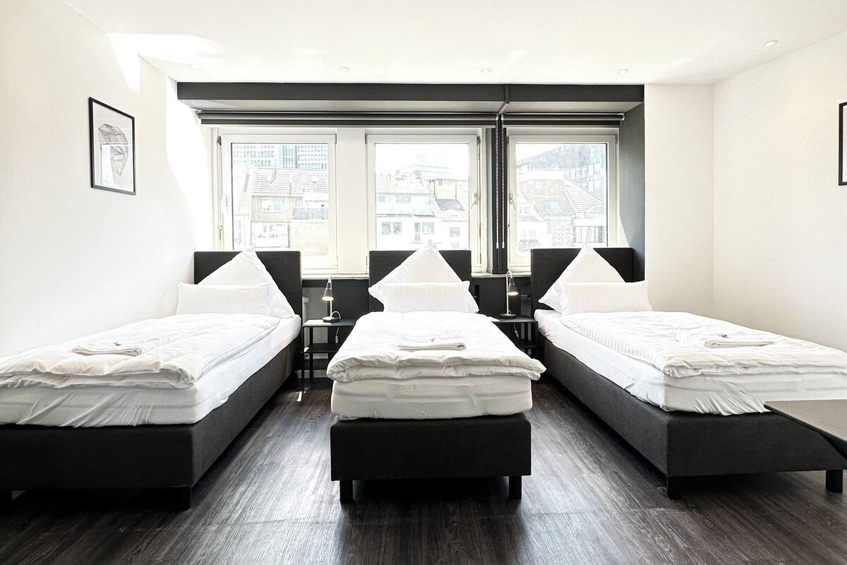A modern hotel room with three single beds, a large window, and a view of the cityscape.