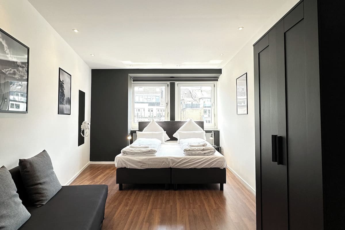 A modern bedroom with a large bed, a dark wardrobe, a sofa, and a window with a city view.