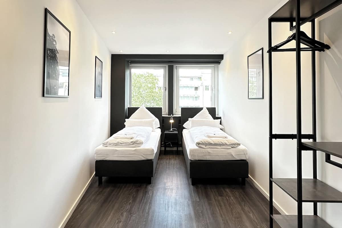 A modern hotel room with two beds, a large window, and a black metal shelf unit.