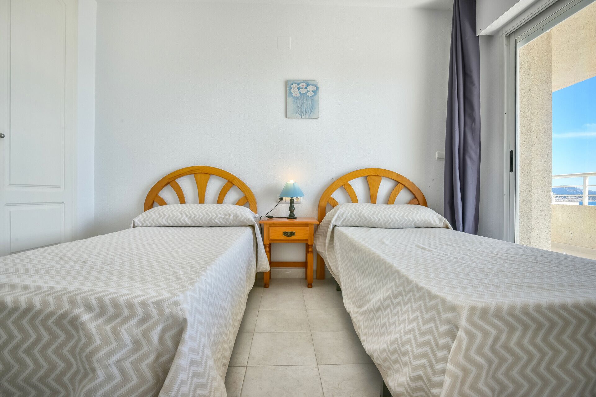 Ein Zimmer mit zwei Einzelbetten, einem Nachttisch mit Lampe, einem kleinen Gemälde an der Wand und Blick durch ein Fenster.