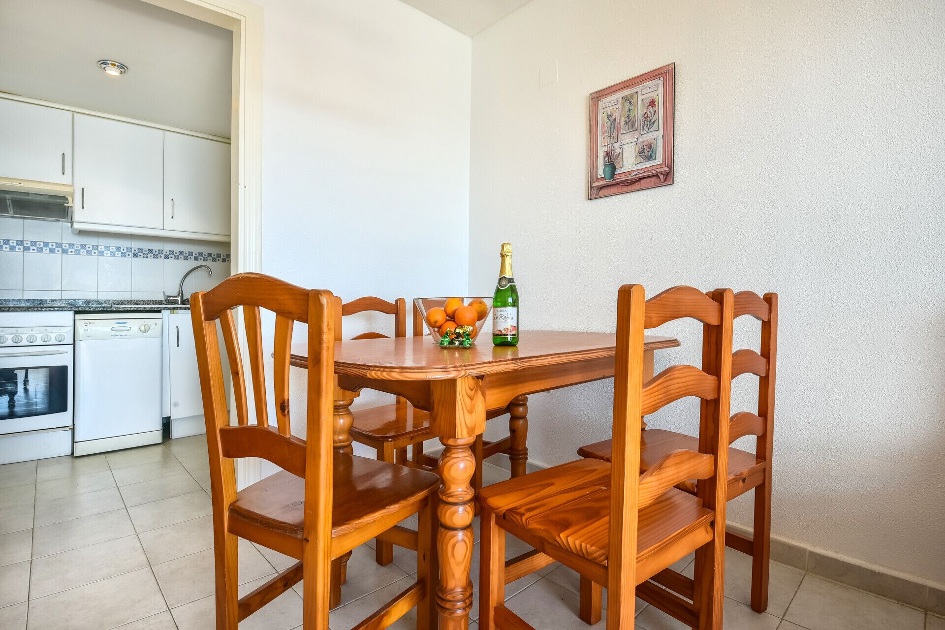 Une table à manger avec des chaises en bois, une bouteille de vin et un saladier rempli d’oranges, dans une pièce dont le sol est carrelé et les murs sont blancs.